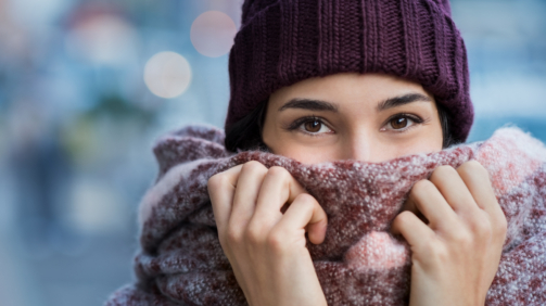 Winter portrait of young beautiful woman covering face with woolen scarf. Closeup of happy girl feeling cold outdoor in the city. Young woman holding scarf and looking at camera."r